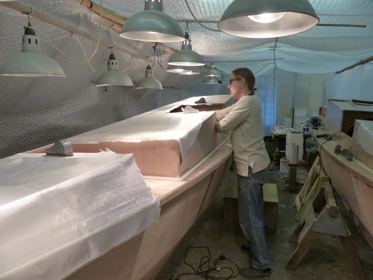 After the glass cloth is cut out on a table, it is laid out on the hull and final trimming is done. The builders used some lead weights to hold it in place. Glass is commonly applied to wooden boats such as Wharram Designs, replacing nylon sheathing used in the past. Compared to mass produced all-GRP boats, however, glass-covered Wharrams utilize considerably less glass. The author is exploring alternative fiber-reinforced composites in their upcoming Wharram Narai Mk IV built on the outer layer of the boat, only to increase durability and the lifespan of the catamaran. (Photo courtesy of Wharram Designs)