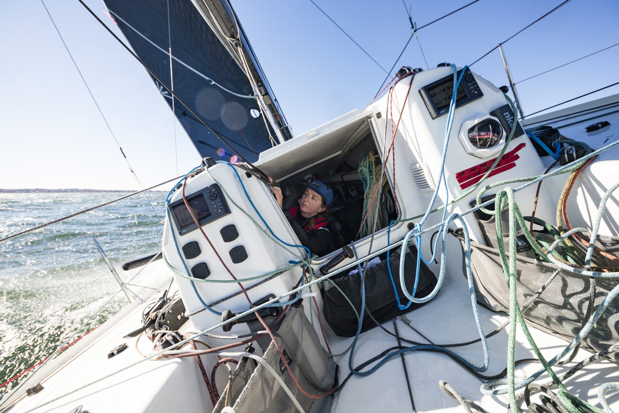 The Figaro 3's cockpit is engineered for solo operation, with dual instrument displays for navigation and weather data, and multiple control lines—including the fractional sloop's mainsail and jib sheets—all within immediate reach of the helmsperson. The boat's polyester sandwich hull with infused PVC foam construction keeps weight to a minimum while maintaining structural integrity. (Photo/ <a href="“https://www.catebrownphoto.com/-/galleries/portfolios/sail”">Cate Brown Photography</a>)
